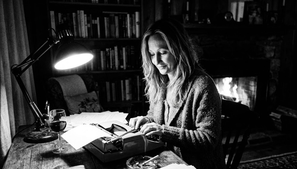 Black-and-white photograph of a woman writing at a typewriter in a study lined with books.